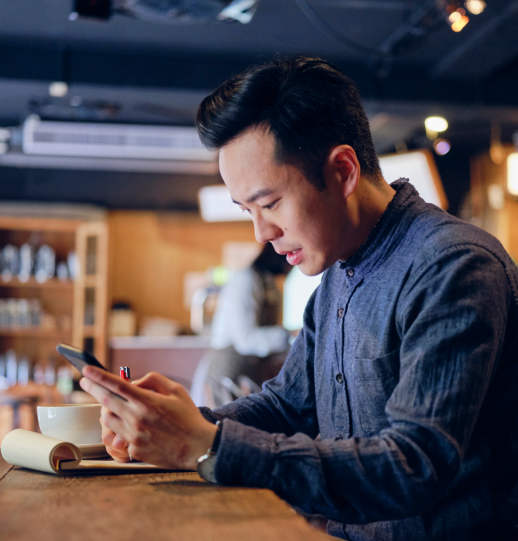 一名男子坐在咖啡店的桌子旁，看着手机并在记事本上写笔记。